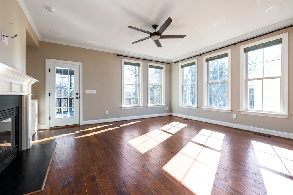 Bright and empty living room with hardwood floor, ceiling fan, and large windows.