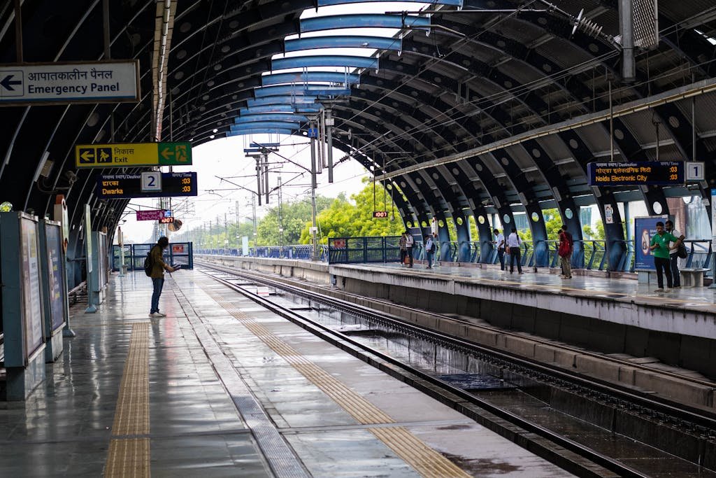 People waiting at a lively New Delhi metro station platform during a rainy day.